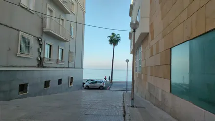Vista del mar desde una calle de Cádiz, entre dos edificios. Al fondo se observa una palmera junto al paseo marítimo, varias personas, vehículos estacionados y un cielo azul despejado.