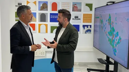 Fran González y José Javier Ruiz Arana conversan de pie en una sala del Rota Tourist Hub. Uno de ellos gesticula con las manos mientras el otro escucha. A su lado hay una gran pantalla con un mapa lleno de puntos de colores y, al fondo, varios paneles decorativos con el logotipo de La Plaza.