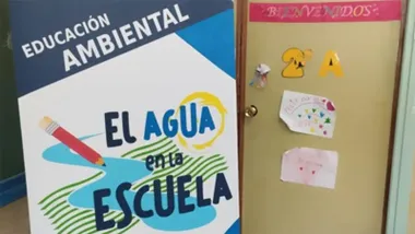 Cartel de "El Agua en la Escuela" con dibujos de agua y un lápiz, junto a una puerta escolar decorada con carteles infantiles de bienvenida.