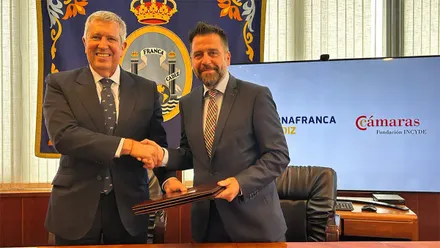 Fran González y Javier Collado, vestidos con traje, se estrechan la mano mientras sostienen una carpeta con el acuerdo firmado. Detrás aparecen el emblema de la Zona Franca de Cádiz y una pantalla con los logotipos de Zona Franca y Fundación INCYDE.