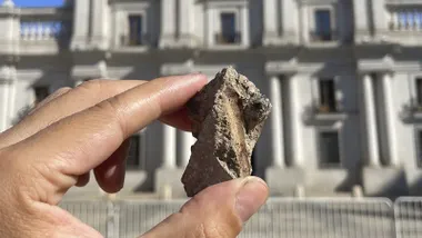 Una mano sostiene un pequeño fragmento de escombro frente a la fachada del Palacio de La Moneda, en Santiago de Chile. El edificio aparece desenfocado al fondo, con una bandera chilena ondeando sobre el tejado y un cielo azul despejado.