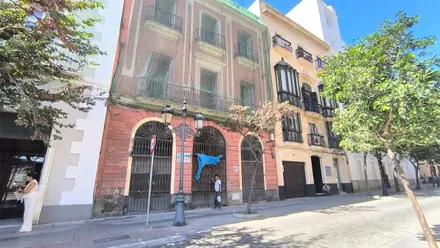 Fachada de un edificio de color rojizo con tres grandes puertas enrejadas en la planta baja, balcones en las plantas superiores y una red de protección que cubre parte del inmueble, situado en el centro de Cádiz.