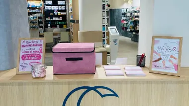 Mostrador de un establecimiento con una urna rosa para depositar mensajes de ánimo contra el cáncer de mama. A su alrededor hay tarjetas, un recipiente con pulseras rosas y dos carteles informativos de la campaña "El Hilo Rosa".