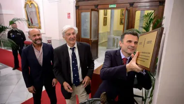 Bruno García coloca una placa dorada del Meridiano de Cádiz junto a la entrada del Salón de Plenos del Ayuntamiento, mientras Miguel Ramos y Juan José Ortiz observan el momento.