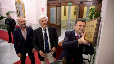 Bruno García coloca una placa dorada del Meridiano de Cádiz junto a la entrada del Salón de Plenos del Ayuntamiento, mientras Miguel Ramos y Juan José Ortiz observan el momento.