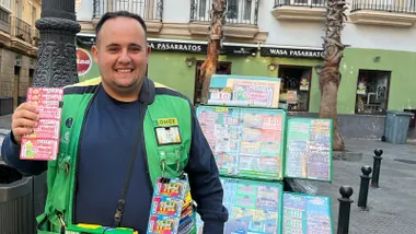 Jesús Batista sonríe mientras muestra varios cupones de la ONCE junto a su puesto de venta en la plaza Antonio Martín de Cádiz. Viste un chaleco verde de la organización y, al fondo, se ven edificios, palmeras y establecimientos comerciales.