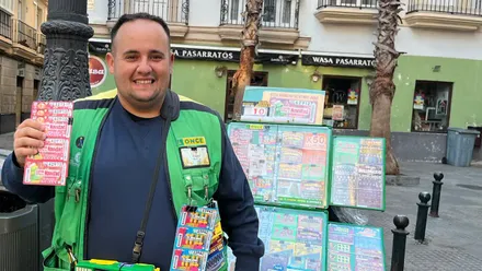 Jesús Batista sonríe mientras muestra varios cupones de la ONCE junto a su puesto de venta en la plaza Antonio Martín de Cádiz. Viste un chaleco verde de la organización y, al fondo, se ven edificios, palmeras y establecimientos comerciales.