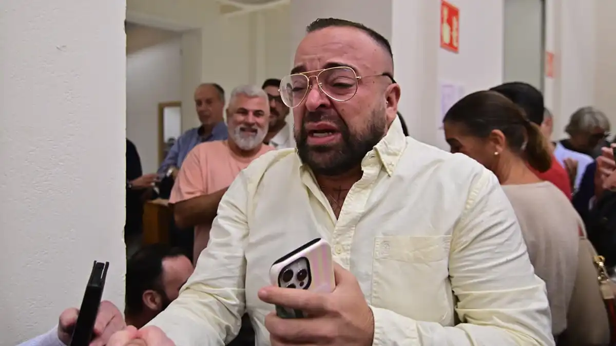 Jesús Macías Graván, con camisa clara y gafas, sostiene un teléfono móvil y muestra una reacción expresiva durante el anuncio de los nombramientos.
