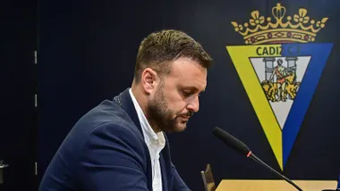 Juan Cala, con chaqueta azul marino y camisa blanca, aparece de perfil ante un micrófono, con la mirada hacia abajo y el escudo del Cádiz CF al fondo.