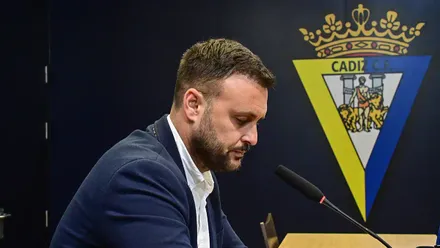 Juan Cala, con chaqueta azul marino y camisa blanca, aparece de perfil ante un micrófono, con la mirada hacia abajo y el escudo del Cádiz CF al fondo.
