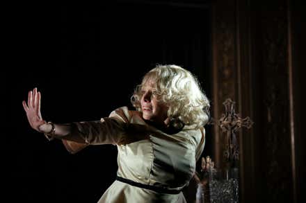 Una intérprete con peluca rubia y vestido claro extiende un brazo con la palma abierta durante una escena teatral, junto a una cruz ornamentada y sobre un escenario de fondo oscuro.
