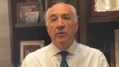 osé Ignacio Landaluce aparece de frente, vestido con camisa blanca y corbata azul, mientras habla a la cámara delante de una estantería con fotografías y documentos enmarcados.