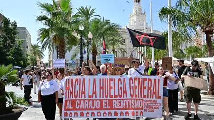 Un grupo de manifestantes marcha por la plaza de San Juan de Dios de Cádiz, frente al Ayuntamiento, sosteniendo una gran pancarta blanca con letras rojas que dice "Hacia la huelga general. Al carajo el rentismo". Detrás se ven más participantes con carteles y banderas.