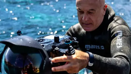 Manu San Félix, vestido con un traje de neopreno negro, sostiene una cámara submarina profesional de grandes dimensiones junto al mar. El explorador observa el equipo mientras lo sujeta con ambas manos, con el agua azul de fondo.