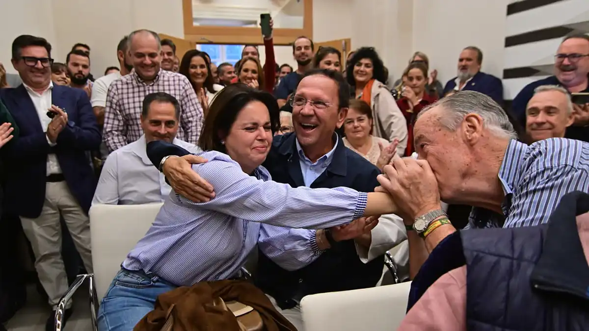 Mari Paz Romero Ruiz recibe un abrazo mientras Isidoro Cárdeno le besa la mano y varias personas aplauden a su alrededor.