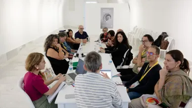 Varias personas participan en una reunión alrededor de una mesa larga en una sala blanca, con ordenadores portátiles, documentos y botellas de agua.