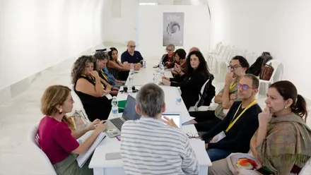 Varias personas participan en una reunión alrededor de una mesa larga en una sala blanca, con ordenadores portátiles, documentos y botellas de agua.