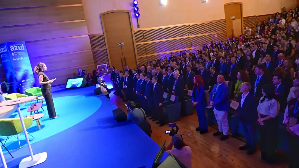 Vista del auditorio del Palacio de Congresos de Cádiz, con el público y los representantes institucionales de pie durante el minuto de silencio. A la izquierda, una presentadora permanece sobre el escenario iluminado en azul, mientras varios fotógrafos captan el momento.