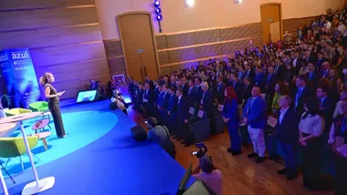 Vista del auditorio del Palacio de Congresos de Cádiz, con el público y los representantes institucionales de pie durante el minuto de silencio. A la izquierda, una presentadora permanece sobre el escenario iluminado en azul, mientras varios fotógrafos captan el momento.
