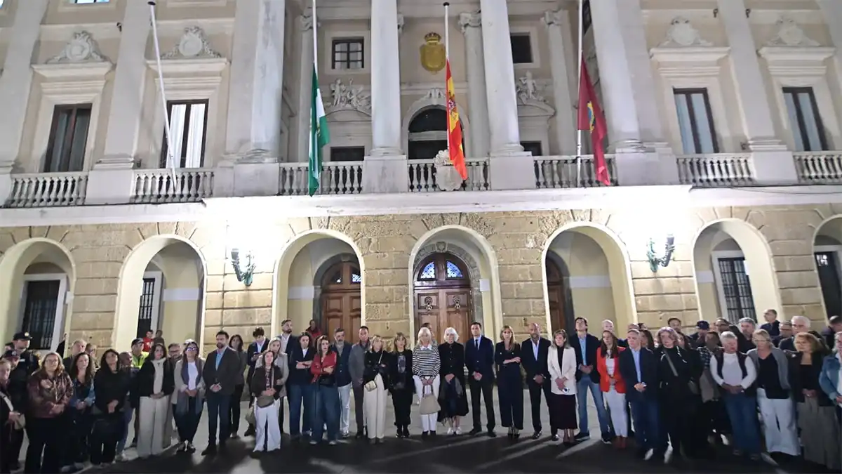 Grupo de ciudadanos y autoridades reunidos de noche ante la fachada iluminada del Ayuntamiento de Cádiz, con las banderas a media asta, durante el homenaje a las víctimas del accidente ferroviario.