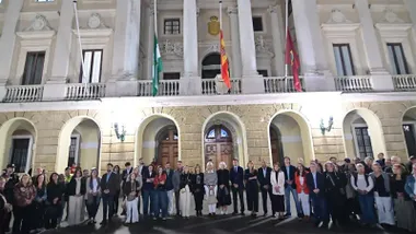 Grupo de ciudadanos y autoridades reunidos de noche ante la fachada iluminada del Ayuntamiento de Cádiz, con las banderas a media asta, durante el homenaje a las víctimas del accidente ferroviario.