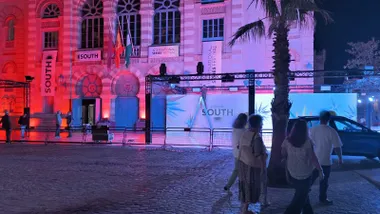 Fachada del Gran Teatro Falla iluminada en rojo y violeta, con carteles del South. Un operario sobre una escalera trabaja en la estructura de iluminación frente al panel del festival, mientras varias personas pasan por la plaza.