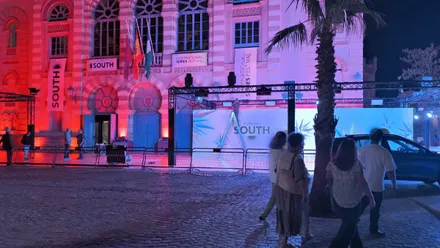 Fachada del Gran Teatro Falla iluminada en rojo y violeta, con carteles del South. Un operario sobre una escalera trabaja en la estructura de iluminación frente al panel del festival, mientras varias personas pasan por la plaza.
