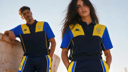 Imagen promocional generada con inteligencia artificial en la que una mujer en primer plano y un hombre al fondo visten polos azul marino, amarillo y azul, con el escudo del Cádiz CF y el logotipo de Kappa, combinados con pantalones deportivos a juego.