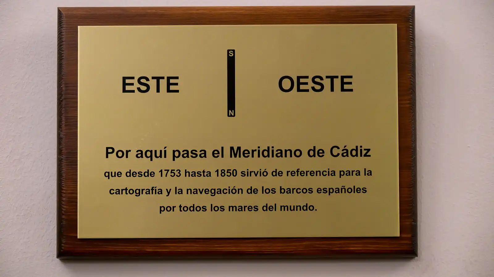 Placa dorada con marco de madera que lleva la inscripción "Por aquí pasa el Meridiano de Cádiz", con las palabras "Este" y "Oeste" separadas por una línea vertical.