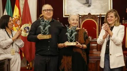 Arístides Vargas y Charo Francés posan sonrientes en el Salón de Plenos del Ayuntamiento de Cádiz, sosteniendo dos esculturas de bronce sobre bases de madera, mientras dos mujeres aplauden a ambos lados.