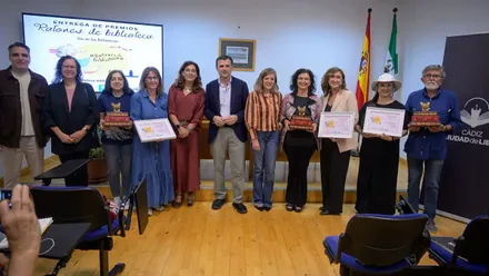 Once personas posan en la Biblioteca Municipal Adolfo Suárez de Cádiz durante la entrega de los Premios Ratones de Biblioteca. Varias sostienen diplomas y trofeos, con el alcalde Bruno García en el centro y un cartel de los premios al fondo.
