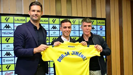 Javier Muiños, a la izquierda, Borja Jiménez, en el centro, y Mario Fuentes, a la derecha, sostienen una camiseta amarilla del Cádiz CF con el nombre del nuevo entrenador, ante un panel de patrocinadores.