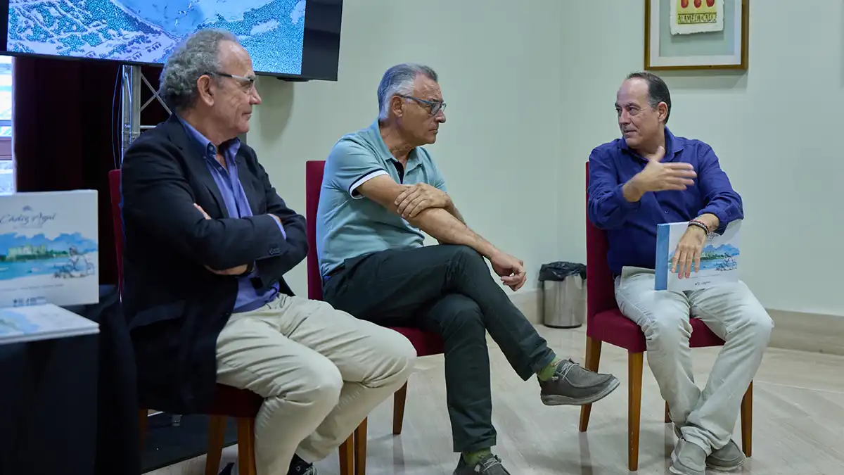 Tres hombres sentados en sillas rojas participan en la presentación del libro "Cádiz Azul: Mar, Tiempo y Paisaje". El hombre de la derecha sostiene un ejemplar y gesticula mientras habla; los otros dos lo escuchan. A la izquierda se observan otros ejemplares de la publicación.