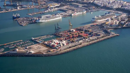 Vista aérea del puerto de Cádiz. En primer plano aparecen la Punta de San Felipe, pequeños pantalanes con embarcaciones y una explanada con contenedores y grúas en el muelle Reina Sofía. El paseo Almirante Pascual Pery bordea el recinto por el lado del mar. Al fondo se ven cruceros atracados y los edificios de la ciudad.