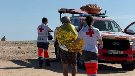 Una persona envuelta en una manta térmica dorada camina junto a una integrante de Cruz Roja hacia un vehículo de rescate. Otro miembro del equipo espera al lado del vehículo, que lleva material de salvamento en el techo. Al fondo se ven el mar y el castillo de Sancti Petri.
