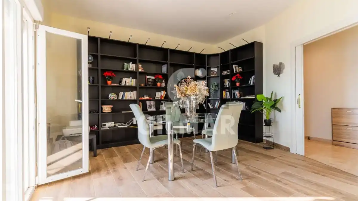 Estancia de la vivienda con suelo de madera, una mesa de cristal con varias sillas y una gran librería negra que ocupa buena parte de la pared del fondo.