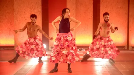 Tres intérpretes sobre un escenario iluminado en tonos rojizos. En el centro, una mujer con camiseta azul oscuro y una voluminosa falda de flores rosas hace un gesto expresivo, mientras dos hombres con el torso descubierto y faldas similares posan a ambos lados.