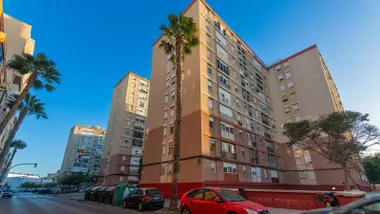 Bloques de viviendas de varias plantas en la barriada de Guillén Moreno de Cádiz, con fachadas de tonos beige y rojizos, palmeras y coches aparcados junto a los edificios.