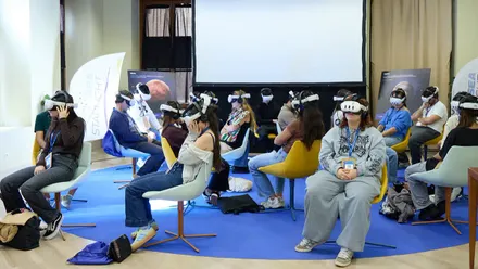 Un grupo de personas sentadas en sillas de distintos colores utiliza gafas de realidad virtual en una sala del Blue Zone Forum. Algunas se ajustan los dispositivos con las manos, mientras otras permanecen observando la experiencia. Al fondo hay una gran pantalla y paneles informativos.